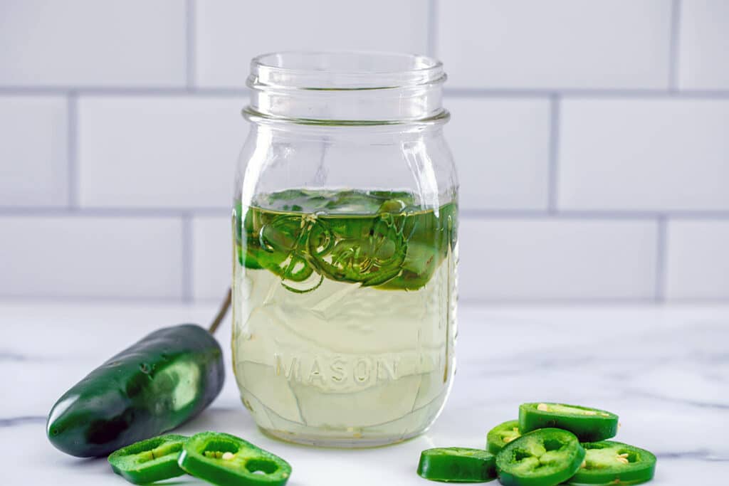 Lanscape head-on view of a mason jar of jalapeño simple syrup with sliced jalapeños and a whole pepper in background