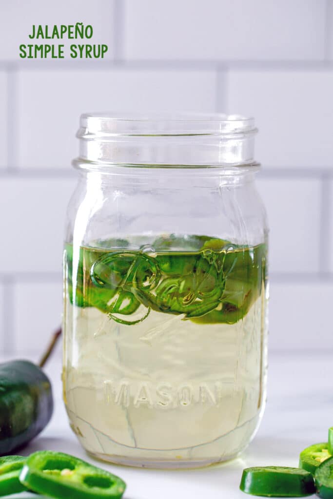 Head-on view of a mason jar filled with jalapeño simple syrup with sliced jalapeños in it and on the table in front with recipe title at top