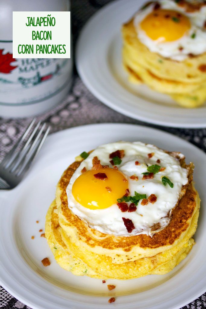 A view from above of a stack of jalapeño bacon corn pancakes with a sunnyside up egg on top on a white plate with a bottle of maple syrup in the background and recipe title at top