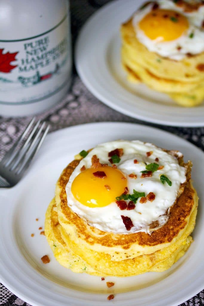 A view from above of a stack of jalapeño bacon corn pancakes with a sunnyside up egg on top on a white plate with a bottle of maple syrup in the background