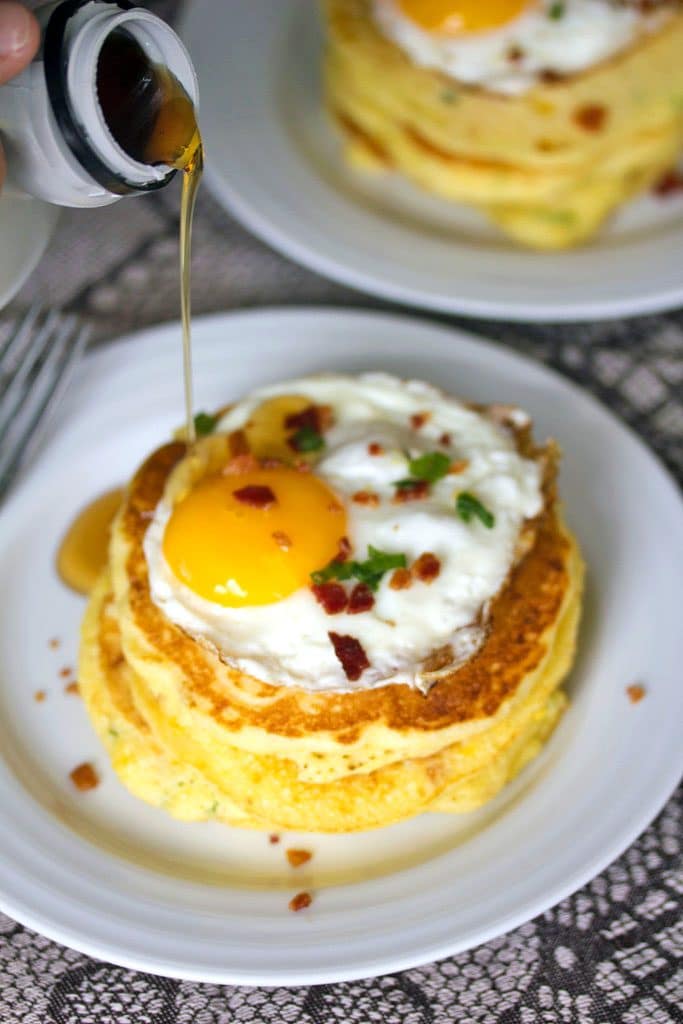 A view from above of a stack of jalapeño bacon corn pancakes with a sunnyside up egg on top on a white plate with maple syrup being poured on them