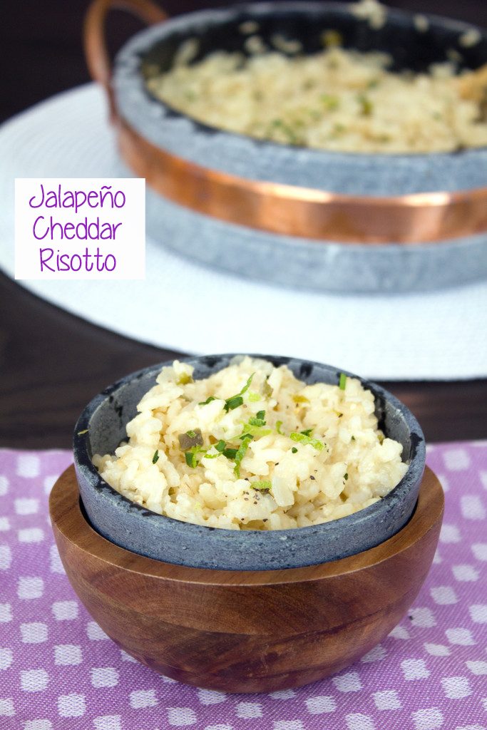 Head-on view of a soapstone bowl of jalapeño cheddar risotto with chopped chives and lime zest on purple placemat with large serving dish of risotto in background and recipe title on side of image