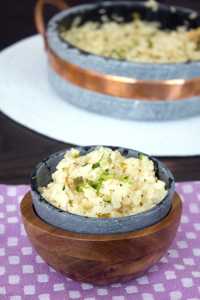 Head-on view of a soapstone bowl of jalapeño cheddar risotto with chopped chives and lime zest on purple placemat with large serving dish of risotto in background