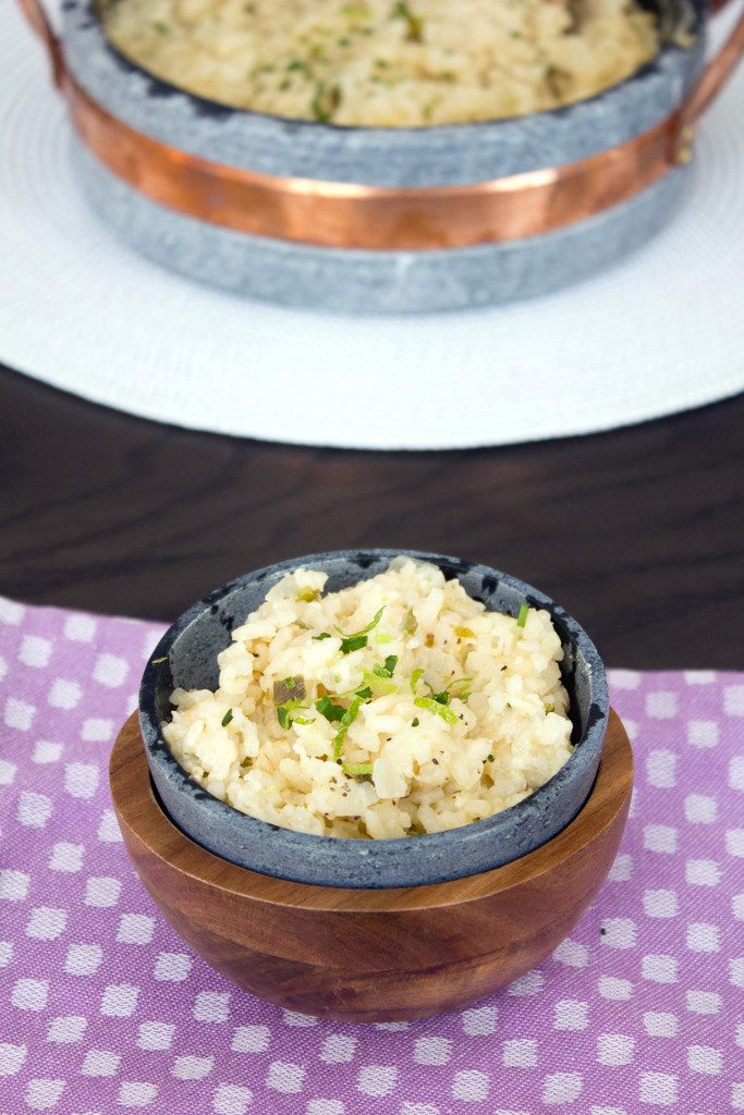 Overhead view of soapstone bowl of jalapeño cheddar risotto topped with chopped chives and lime zest on purple placemat with serving dish of risotto in backrgound