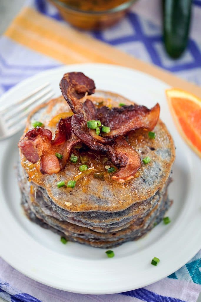 Bird's eye view of jalapeño cornmeal pancakes topped with crispy strips of bacon, diced jalapeños, and orange marmalade syrup on a white plate with orange slice in the background