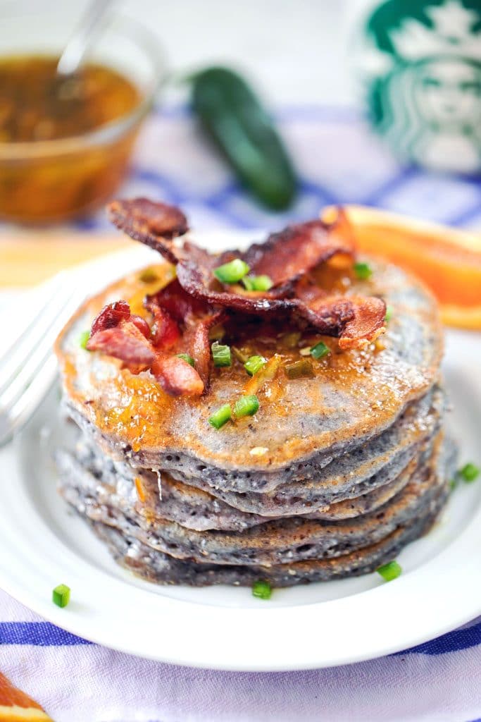 Close-up view of stack of blue jalapeño cornmeal pancakes topped with bacon and diced jalapeño on a white plate with small dish or orange marmalade syrup, jalapeño, and Starbucks mug in the background
