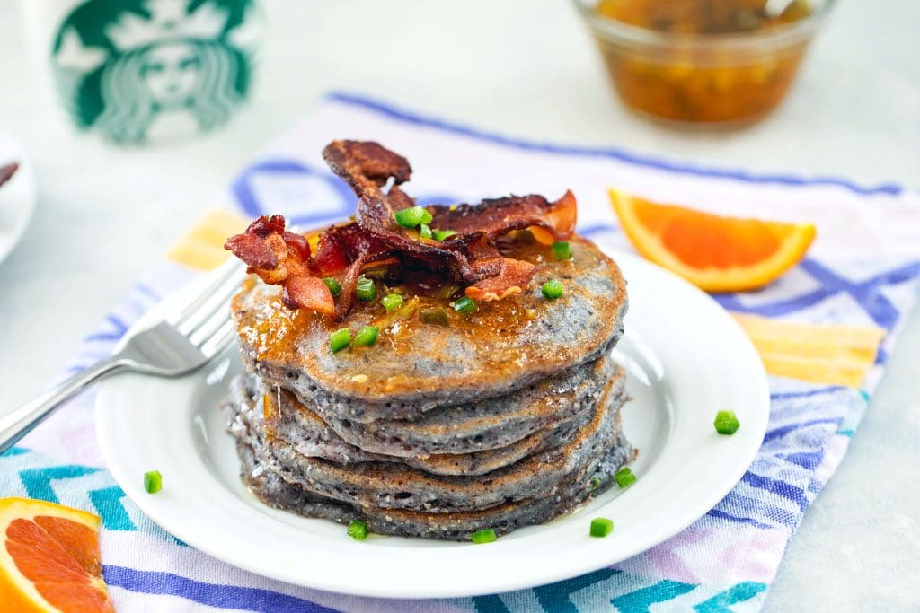 Head on view of stack of blue jalapeño cornmeal pancakes topped with crispy bacon on a white plate and purple and yellow napkin with orange slices and Starbucks mug in the background