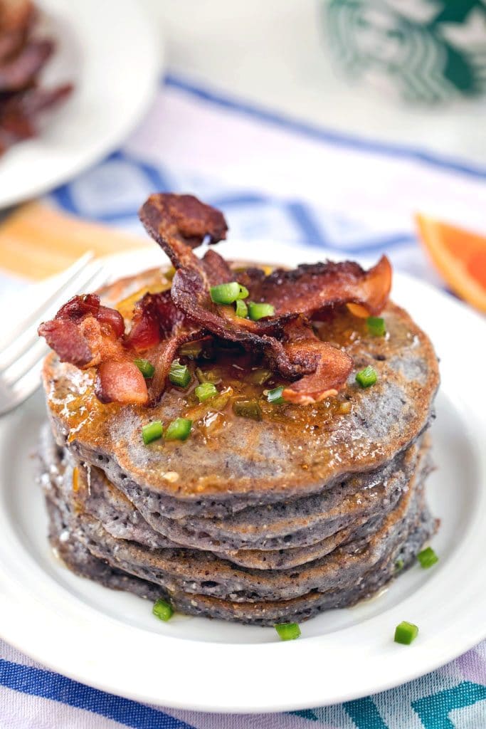 Hero image of blue jalapeño cornmeal pancakes in a large stack, topped with crispy bacon strips, orange syrup, and chopped jalapeño on a white plate