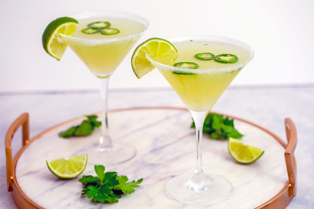 Head-on landscape view of two jalapeño martinis with lime wedge and jalapeño garnish on a marble tray with parsley and lime wedges in background