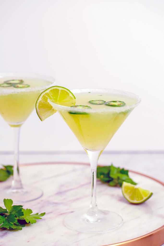 Head-on view of a jalapeño martini on a marble tray with lime wedge, parsley, and second cocktail in the background