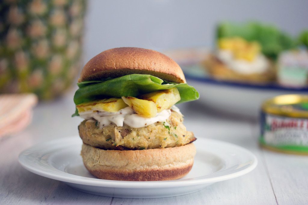 Landscape head-on view of jalapeño tuna burger with grilled pineapple, lemon mayo, and lettuce with second burger, pineapple, and can of tuna in background