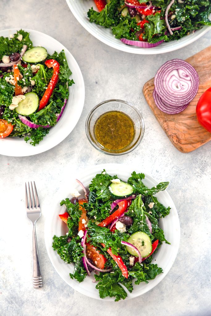 Overhead view from a distance of three bowls of kale Greek salad, fork, bowl of Greek dressing, and cutting board with sliced red onion and tomato