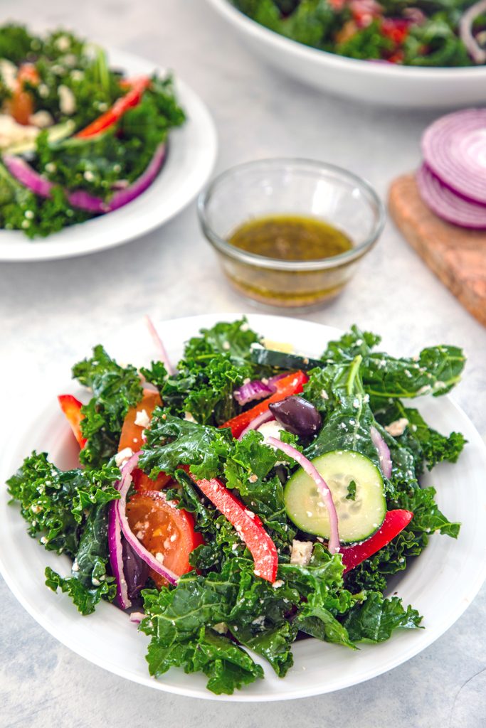 Head-on closeup view of a bowl of kale Greek salad with bowl of dressing, sliced red onion, and two more bowls of salad in background