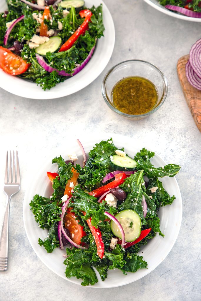 Overhead view of kale Greek salad in a serving bowl with fork, bowl of dressing, and second salad bowl in the background