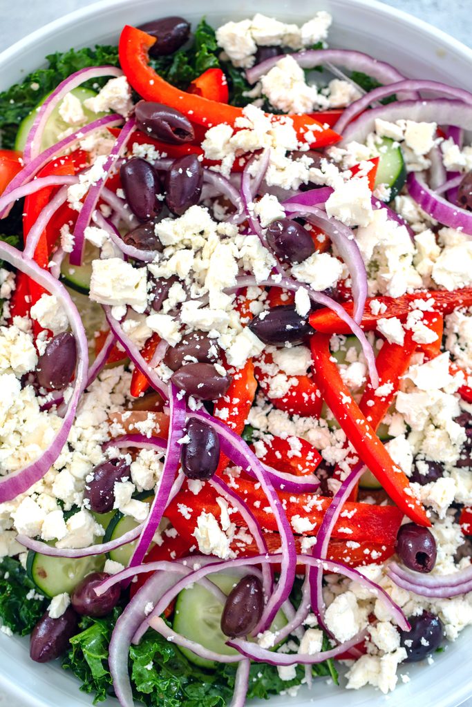 Overhead view of ingredients for kale Greek salad salad in bowl, including kale, cucumber, red pepper, onion, tomato, kalamata olive, and feta