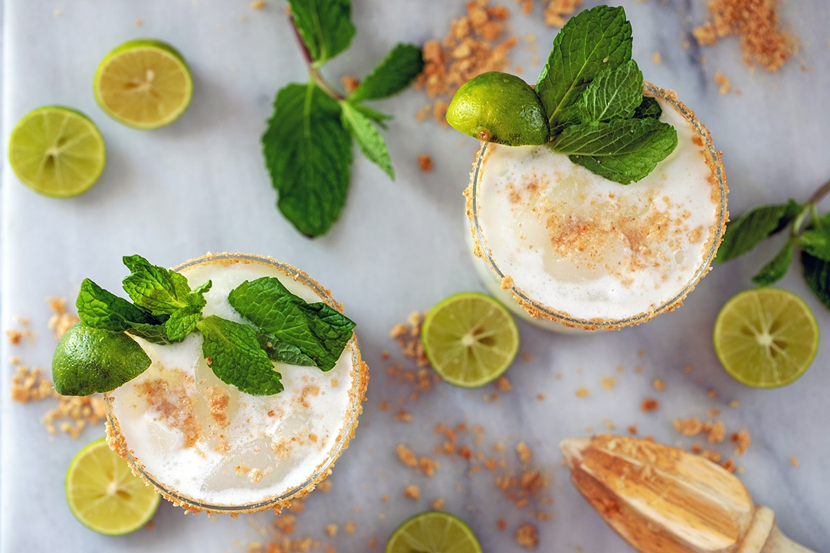 Landscape bird's eye view of two key lime pie mojitos garnished with graham cracker crumbs, key limes, and mint leaves and garnishes scattered around the glasses.