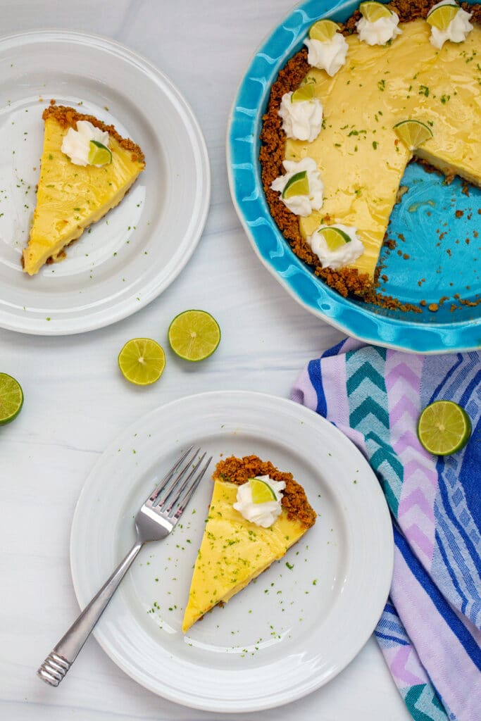 Bird's eye view of slice of key lime pie on plate with second slice in background and remaining pie in pie plate with key lime halves all around