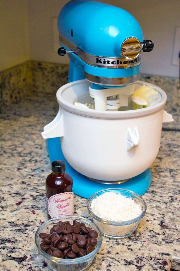 Teal KitchenAid mixer with ice cream attachment running and bowls of dark chocolate chips and coconut and vanilla extract