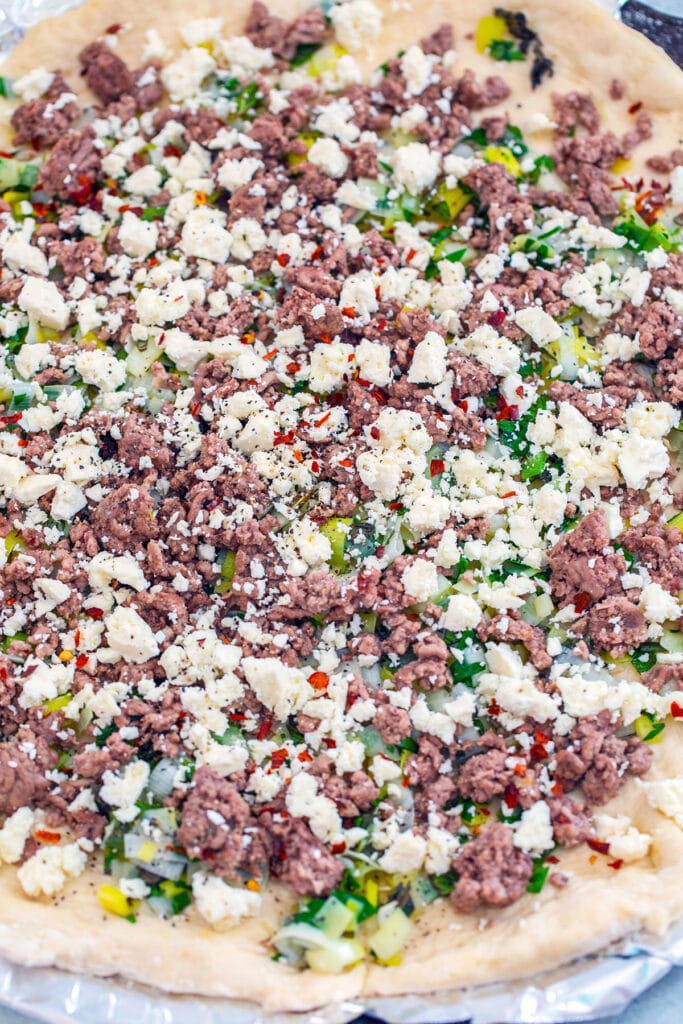 Overhead view of flatbread dough rolled out and topped with ground lamb, crumbled feta, leeks, scallions, mint, and red pepper flakes