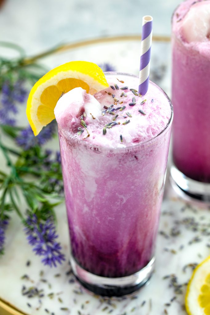 Overhead view of a purple lavender lemonade float in a tall glass with lemon wedge garnish, dried lavender, and striped straw, with lavender and a second drink in the background