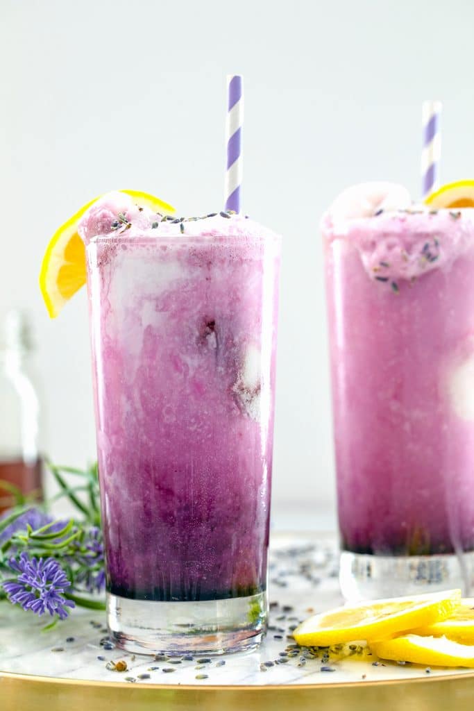 Head-on view of two lavender lemonade floats in tall glasses on a marble surface with lemons and lavender all around