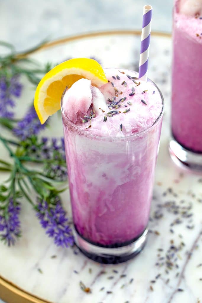 Overhead view of a lavender lemonade float with dried lavender, lemon wedge garnish, and purple and white striped straw on a marble surface with lavender in the background