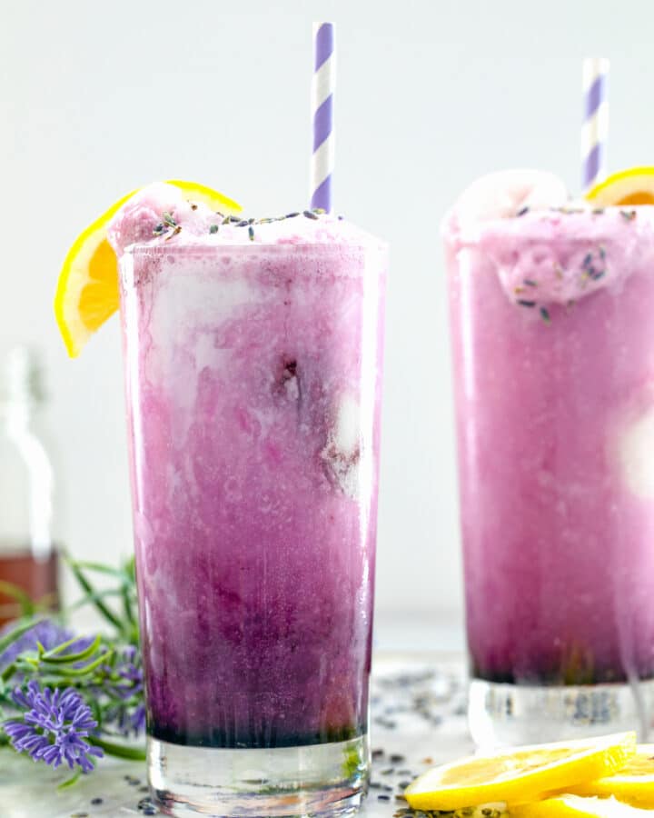 Head-on view of two tall glasses of bright purple lavender lemonade floats with lemons and straws