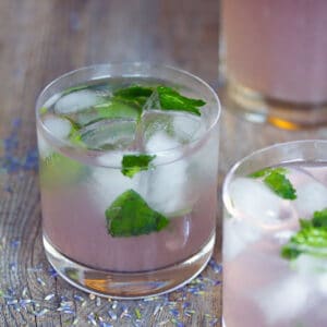 Two lavender lemonade mojitos with ice and mint on wooden surface with lavender buds all around