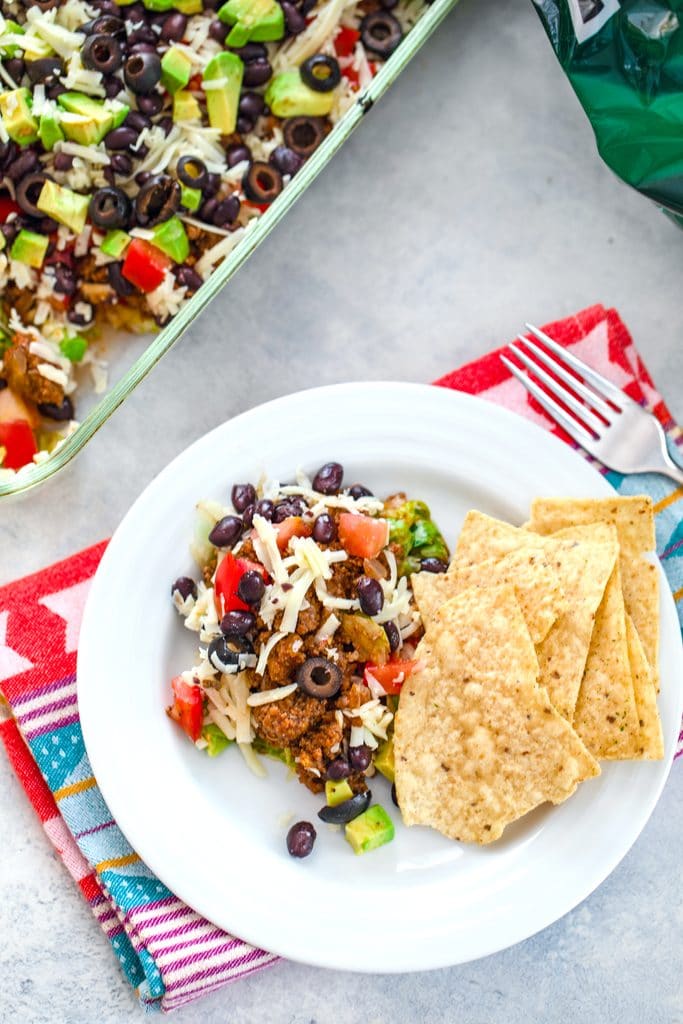 Overhead view of layered taco salad serving on a white plate with chips, along with baking dish of salad in the background, fork, and bag of chips