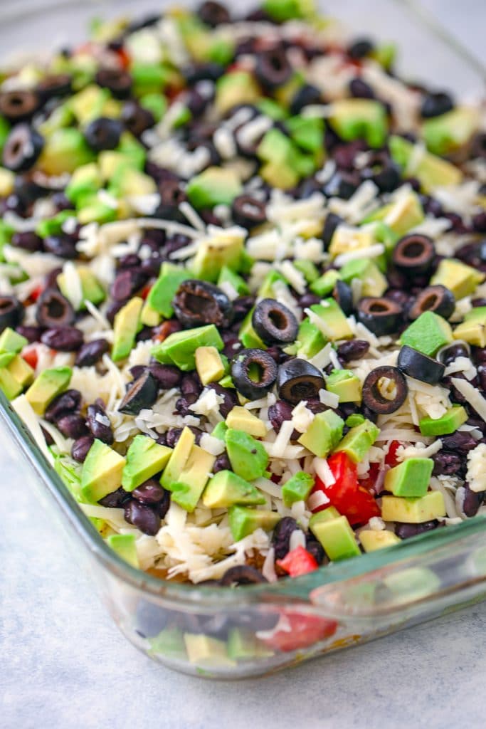 Complete layered taco salad in baking dish with lettuce, beef, cheese, black beans, tomatoes, avocado, and olives