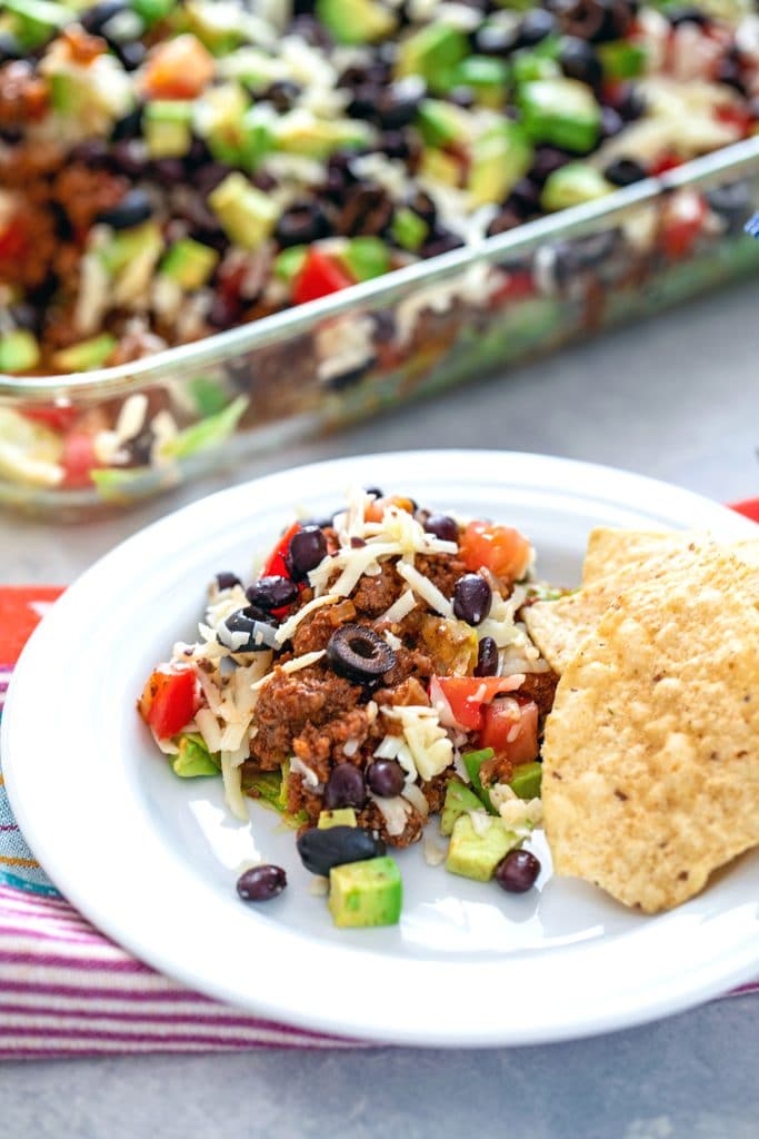 Head-on view of layered taco salad serving on a white plate with tortilla chips with the rest of the dish in the background