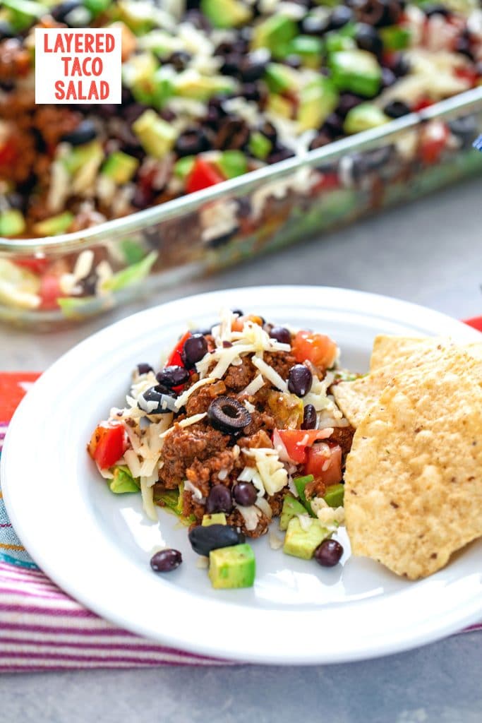 Head-on view of layered taco salad serving on a white plate with tortilla chips with the rest of the dish in the background and "Layered Taco Salad" text at top