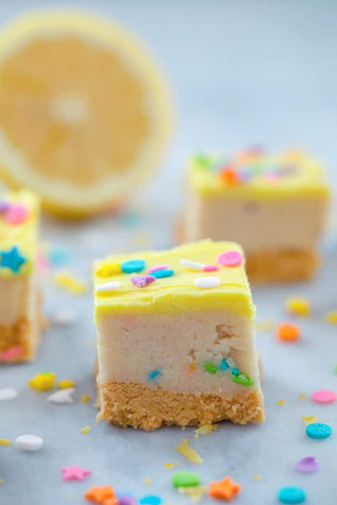 One lemon cookie dough bar sitting on a grey table with other cookie dough squares and a lemon half in the background and sprinkles scattered around