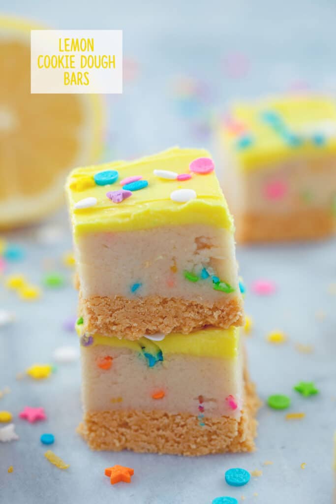 Head-on view of two lemon cookie dough bars stacked on each other with sprinkles and a lemon half in the background and "Lemon Cookie Dough Bars" text at top