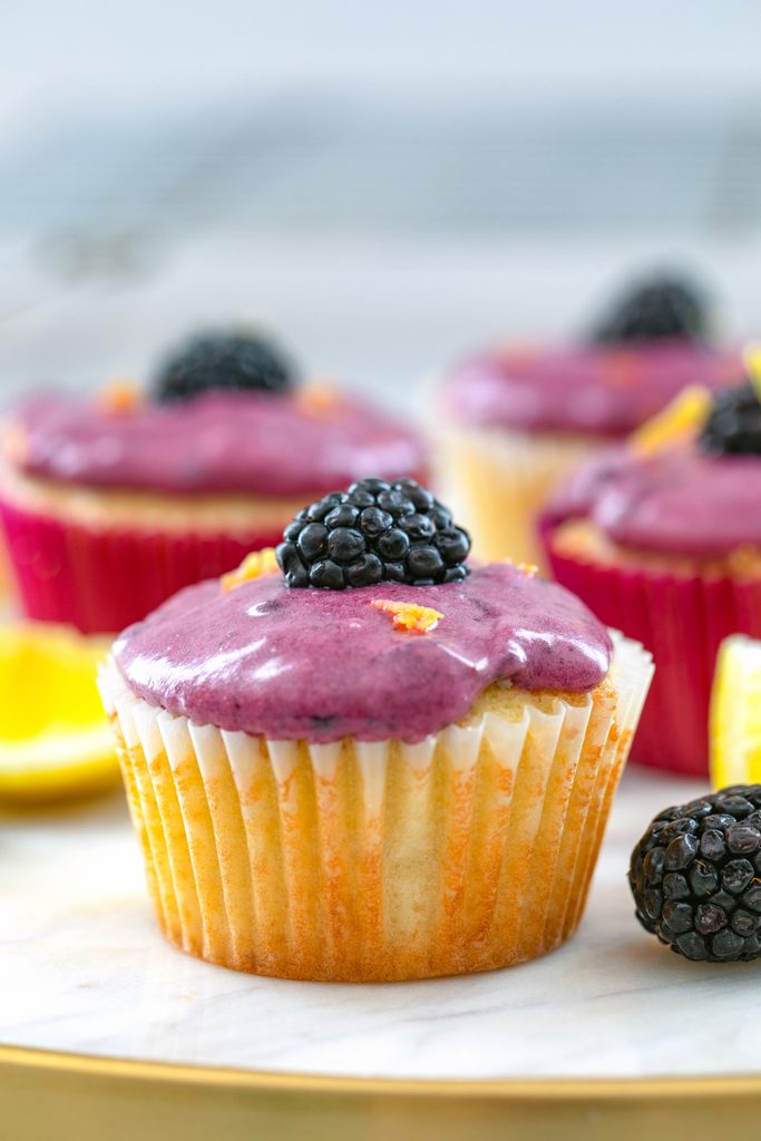 Head-on view of a lemon cupcake with blackberry cream cheese frosting topped with a blackberry on a marble tray with more cupcakes, blackberries, and lemon wedges in the background