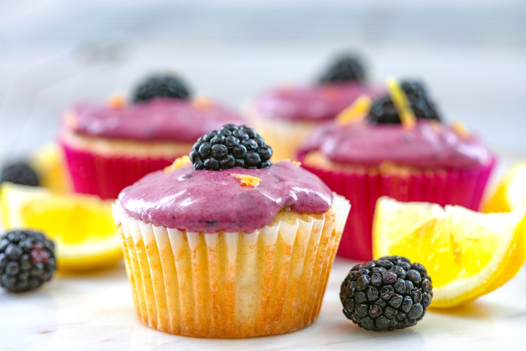 Landscape head-on view of lemon cupcake with blackberry cream cheese frosting topped with blackberry with blackberries with more cupcakes and lemon wedges in the background