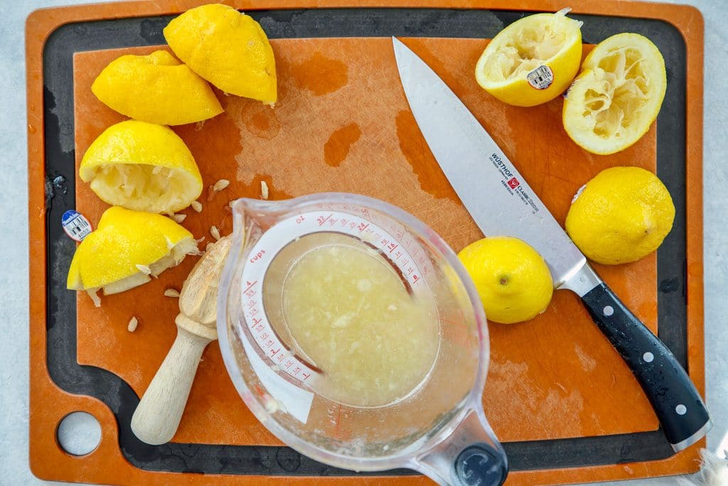 Wood cutting board with juiced lemon halves, a knife, a citrus reamer, and a measuring cup filled with lemon juice