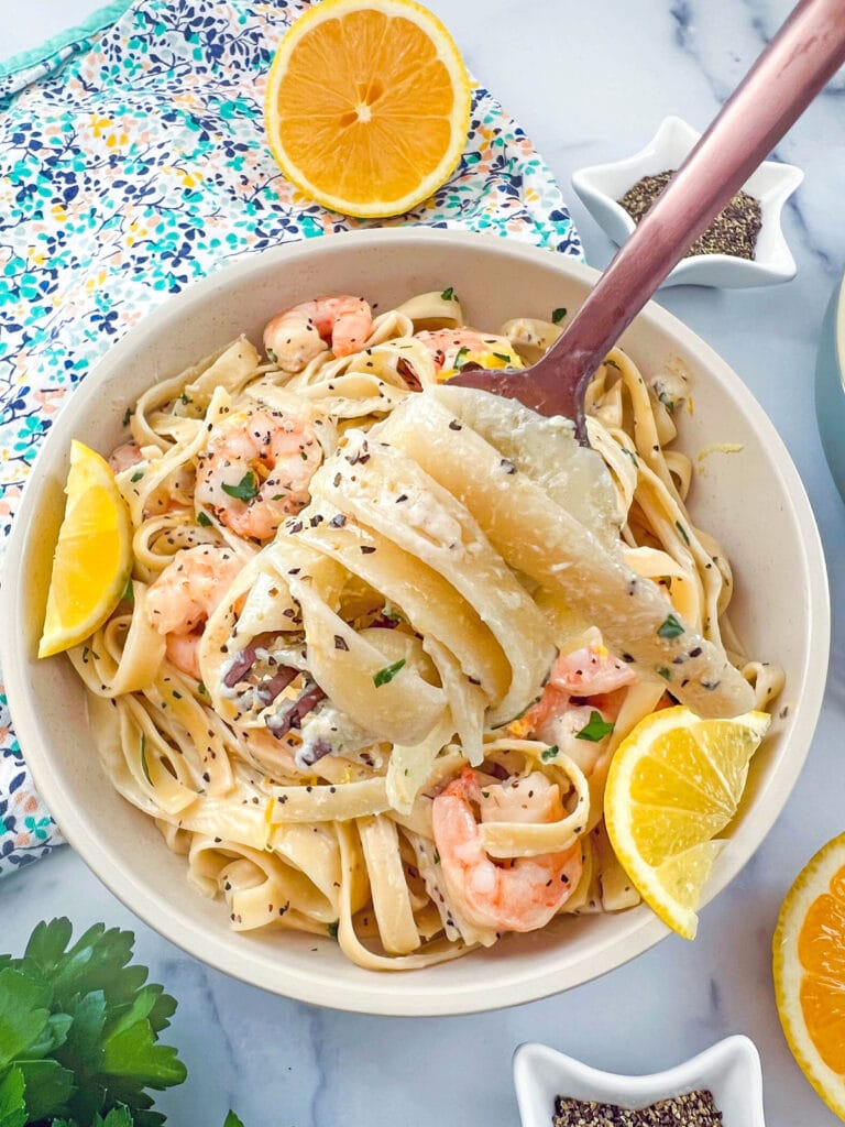 Fork holding up twirled linguine with lemon pepper cream sauce over bowl of pasta with shrimp and lemon.