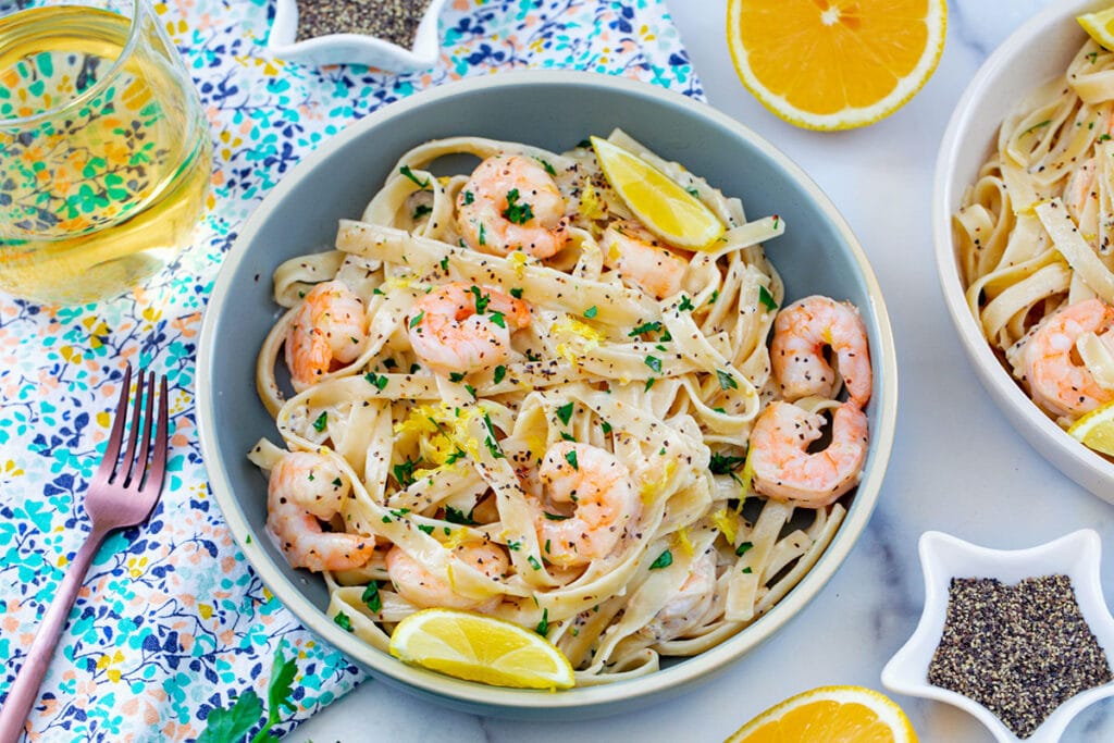 Landscape overhead view of lemon pepper pasta with shrimp and lemon wedges with rose gold fork and star-shaped bowls filled with pepper in background.