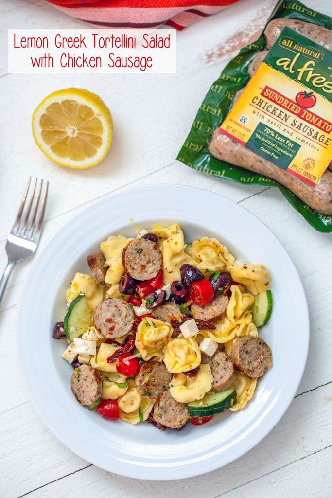 Overhead view of white bowl with lemon greek tortellini salad with fork, lemon half, and sundried tomato chicken sausage package in the background with "Lemon Greek Tortellini Salad with Chicken Sausage" text at top