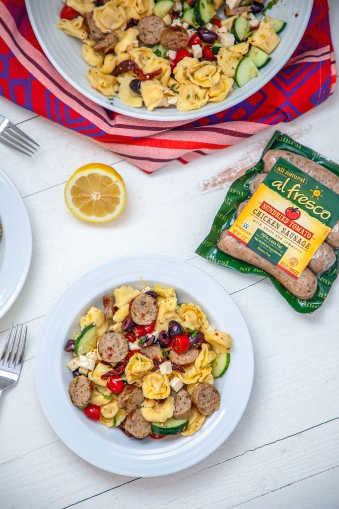 Overhead from a distance view of white serving bowl of tortellini salad with serving bowl, chicken sausage package, lemon half, and forks in the background