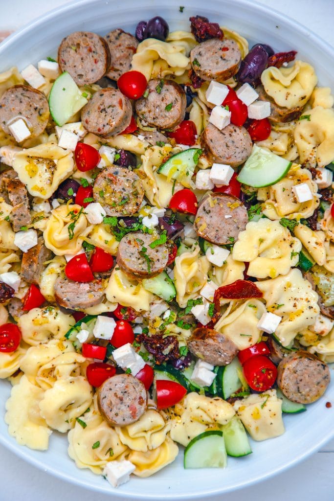 Overhead close-up of lemon Greek tortellini salad in a white bowl with bright colors from tomatoes, cucumber, feta, lemon, and mint