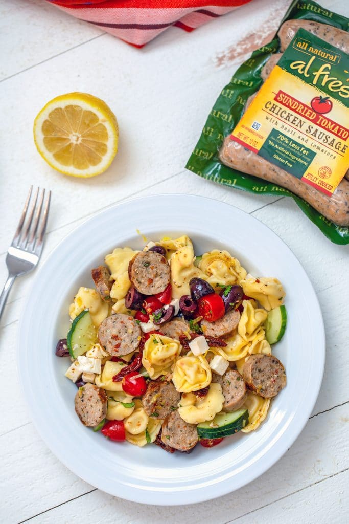Overhead view of white bowl with lemon greek tortellini salad with fork, lemon half, and sundried tomato chicken sausage package in the background