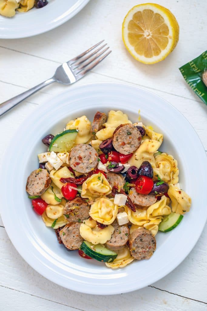 Overhead close-up view of white serving bowl with lemon greek tortellini salad with fork and lemon half in the background