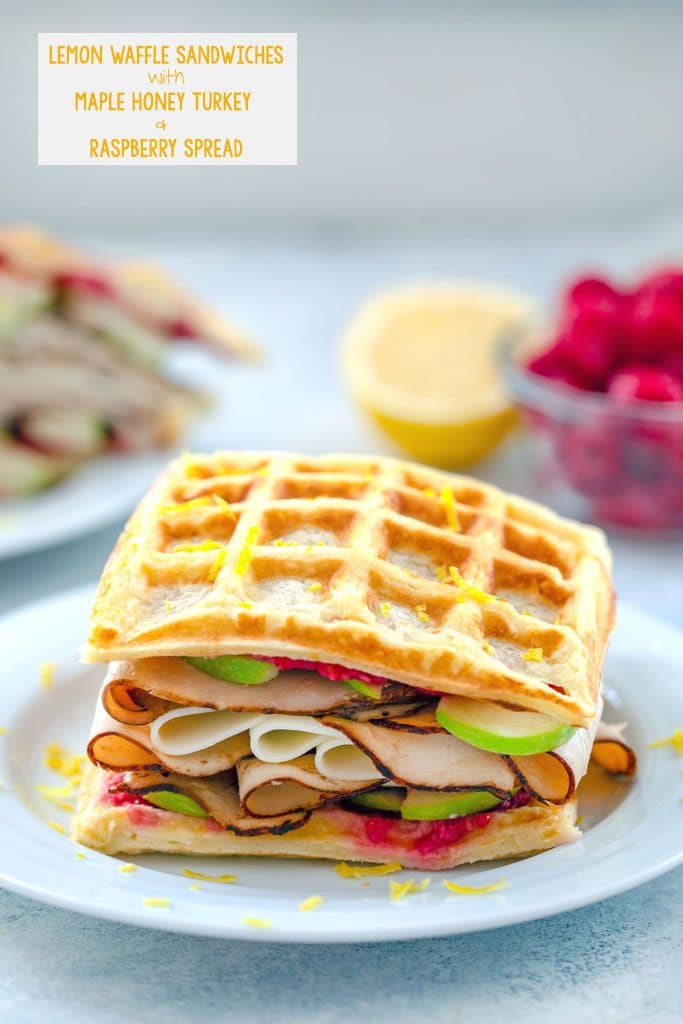 Head-on view of lemon waffle sandwiches loaded with turkey, cheese, raspberry spread, and green apples with raspberries and lemon half in background and recipe title text at top