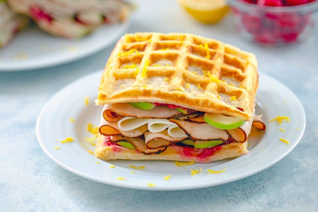 Landscape view of lemon waffle sandwich on a white plate loaded with turkey, cheese, green apples, and raspberry spread with lemon zest sprinkled on top