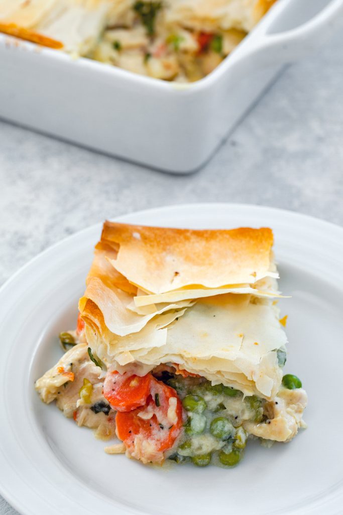 Overhead view of a serving of lighter chicken pot pie with peas and carrots on a white plate with the baking dish in the background