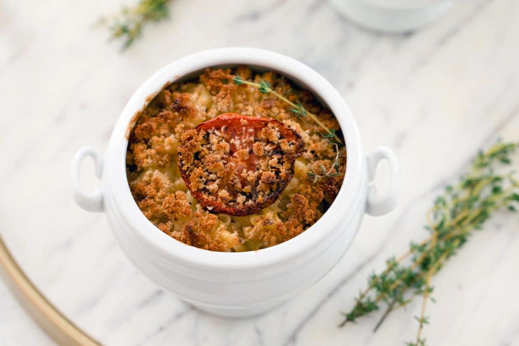 Overhead landscape view of a white ramekin of lighter mac and cheese with roasted tomatoes and breadcrumbs on a marble tray with thyme sprigs all around