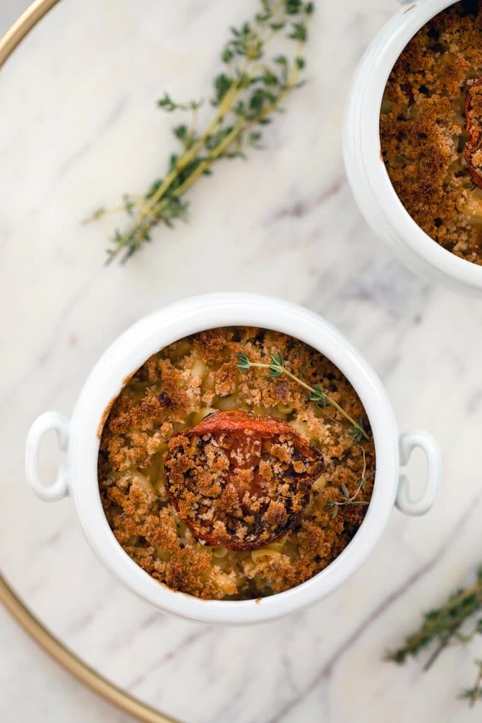 Bird's eye view of two white ramekins of lighter macaroni and cheese with roasted tomatoes and breadcrumbs on a marble tray with thyme