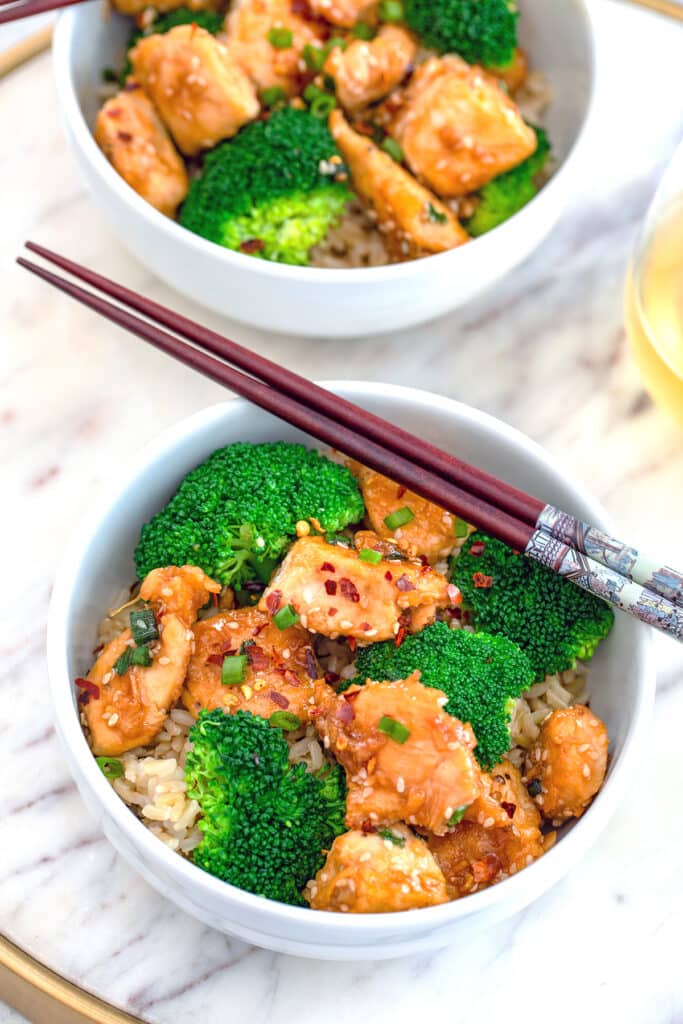 Overhead view of a white bowl of sesame chicken and broccoli over rice in a white bowl with chopsticks on a marble surface with a second bowl in the background.